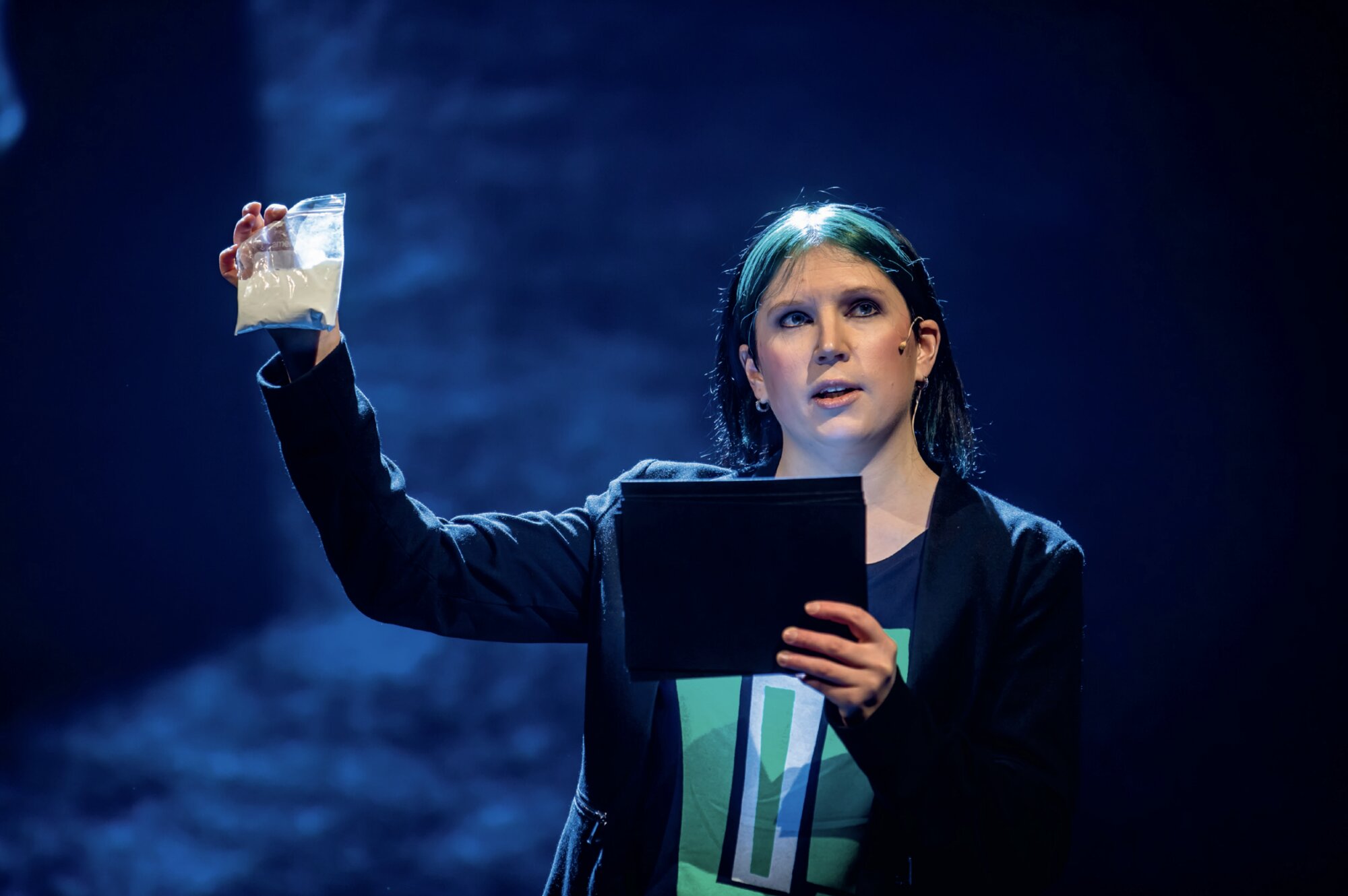 Anni Pasanen on a blue-lit stage holding up a bag of white caffeine powder while reading from a folder