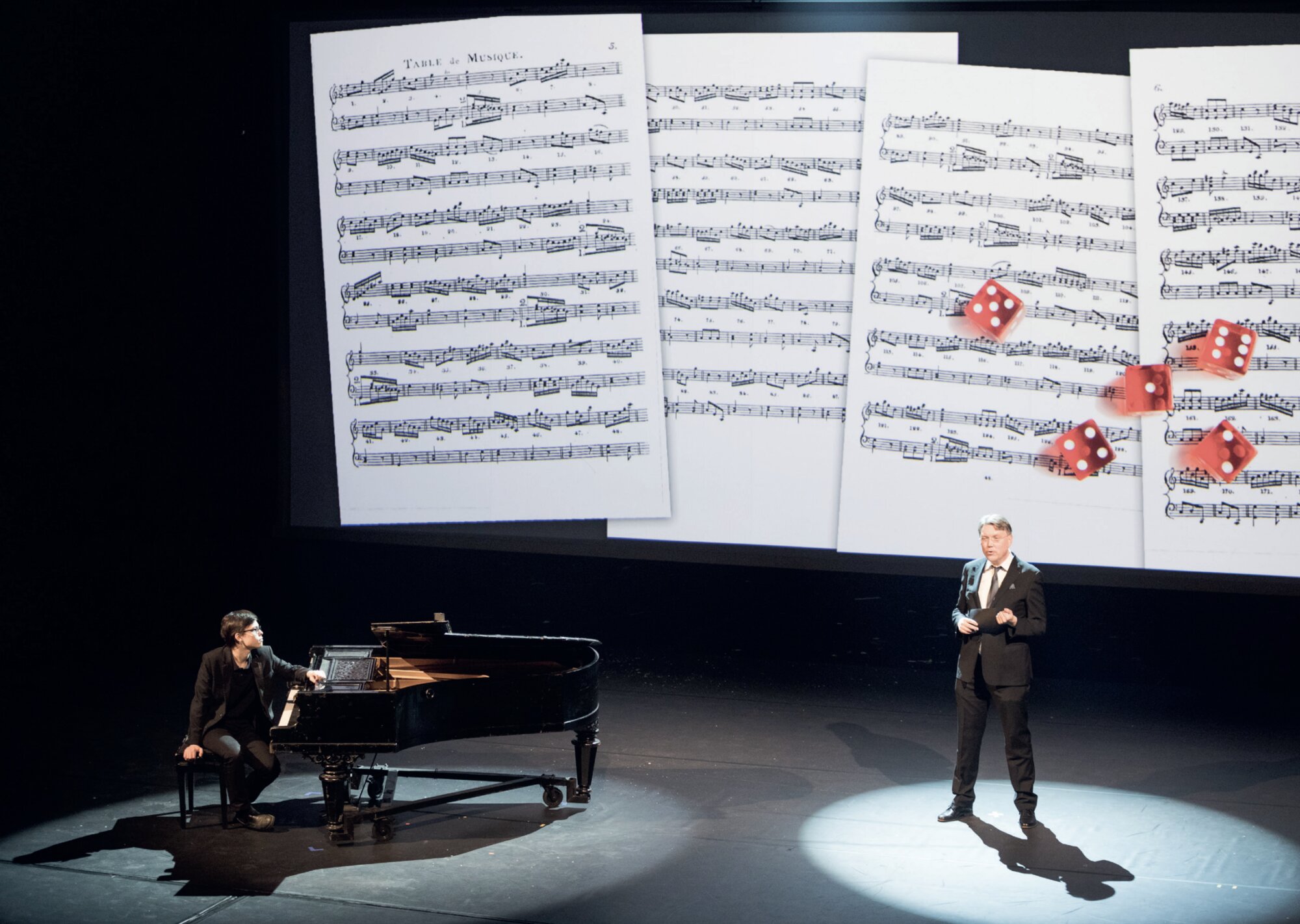 Vesa Sirén speaking on stage as a pianist plays, with sheet music and red dice projected behind them
