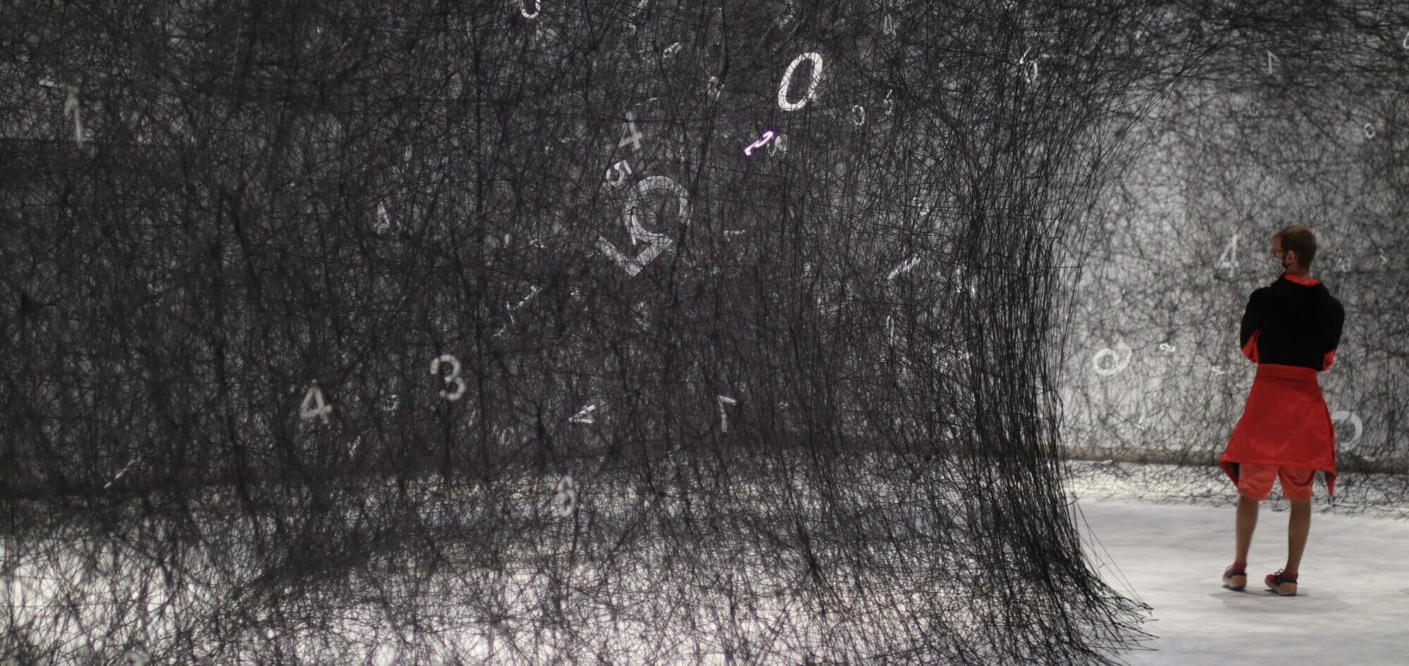 Person in a red skirt standing in an immersive installation of hanging black threads woven with scattered white numbers