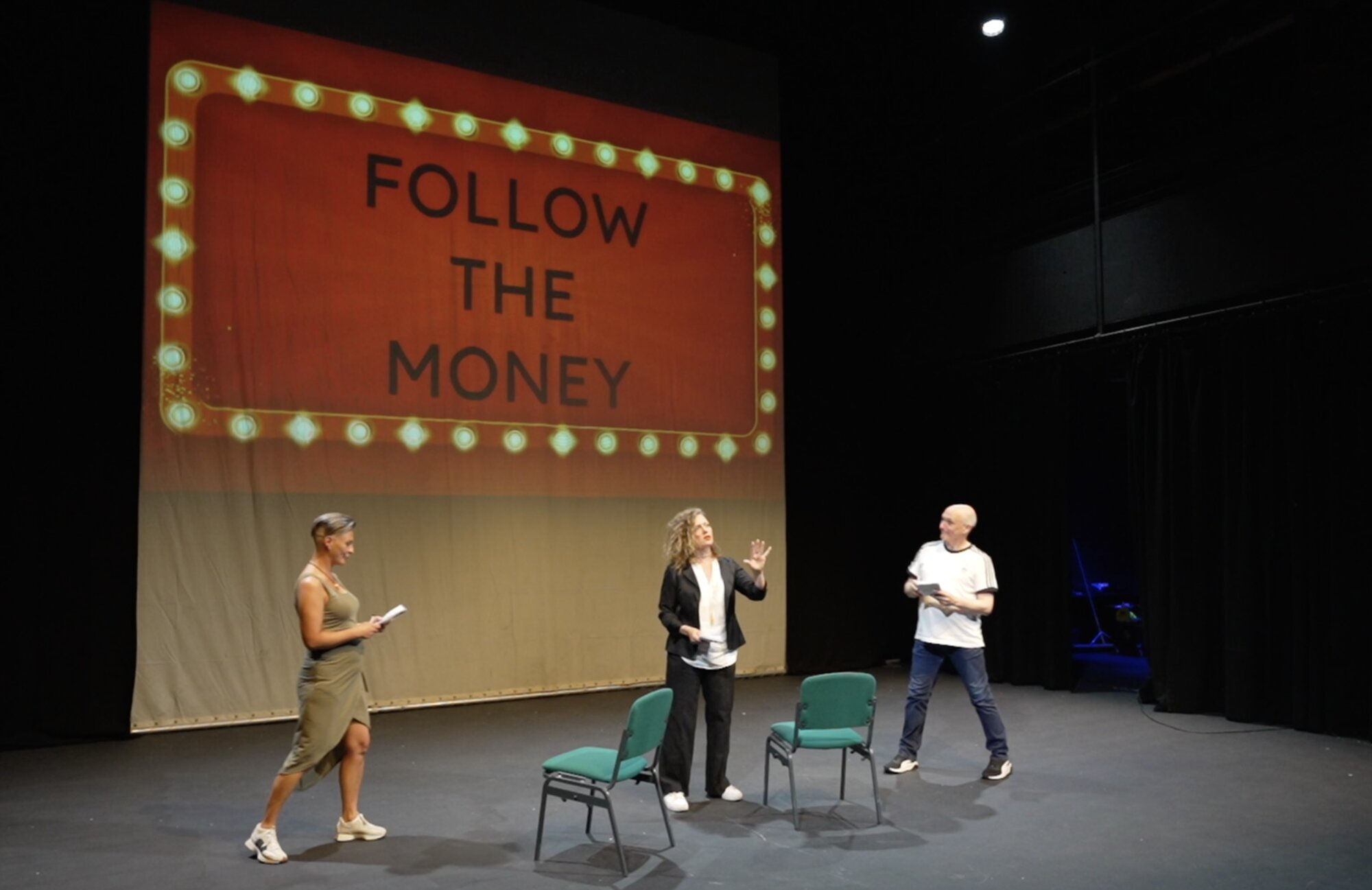 Three performers act out a gameshow scene by two empty chairs, under a projected 'Follow the Money' marquee sign