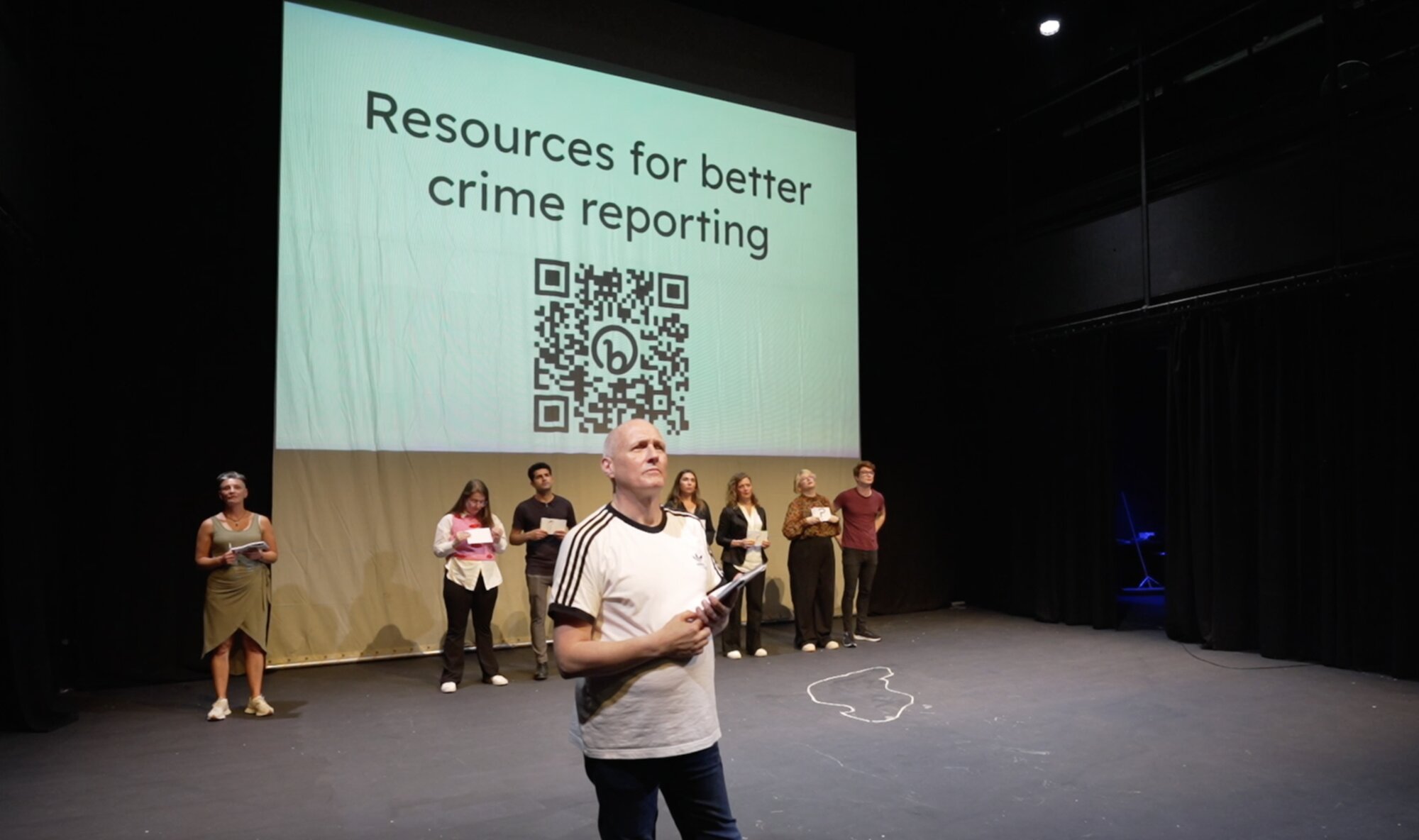 A performer addresses the audience as cast members stand behind, under a projected QR code for 'Resources for better crime reporting'