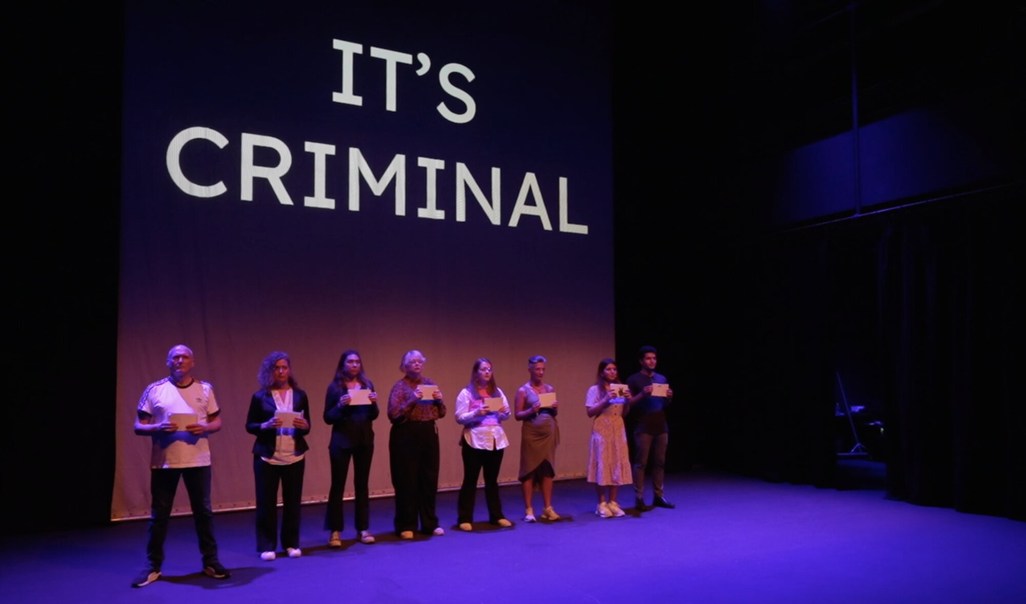 Eight performers stand in a line on stage holding cards beneath a large projected 'It's Criminal' title