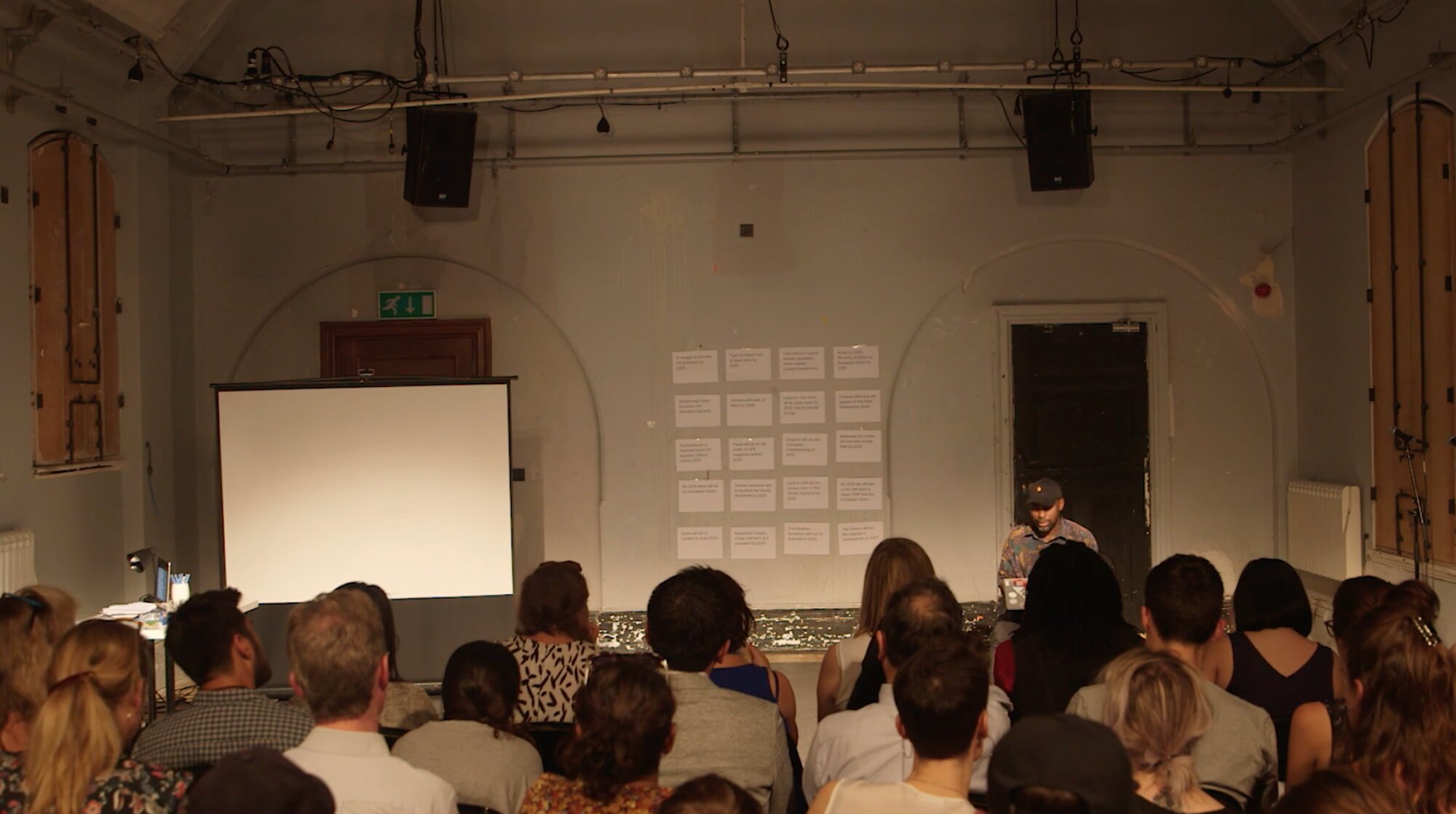 Seated audience watching a presenter beside a grid of question cards taped to the wall for Harun Morrison's prediction-market piece