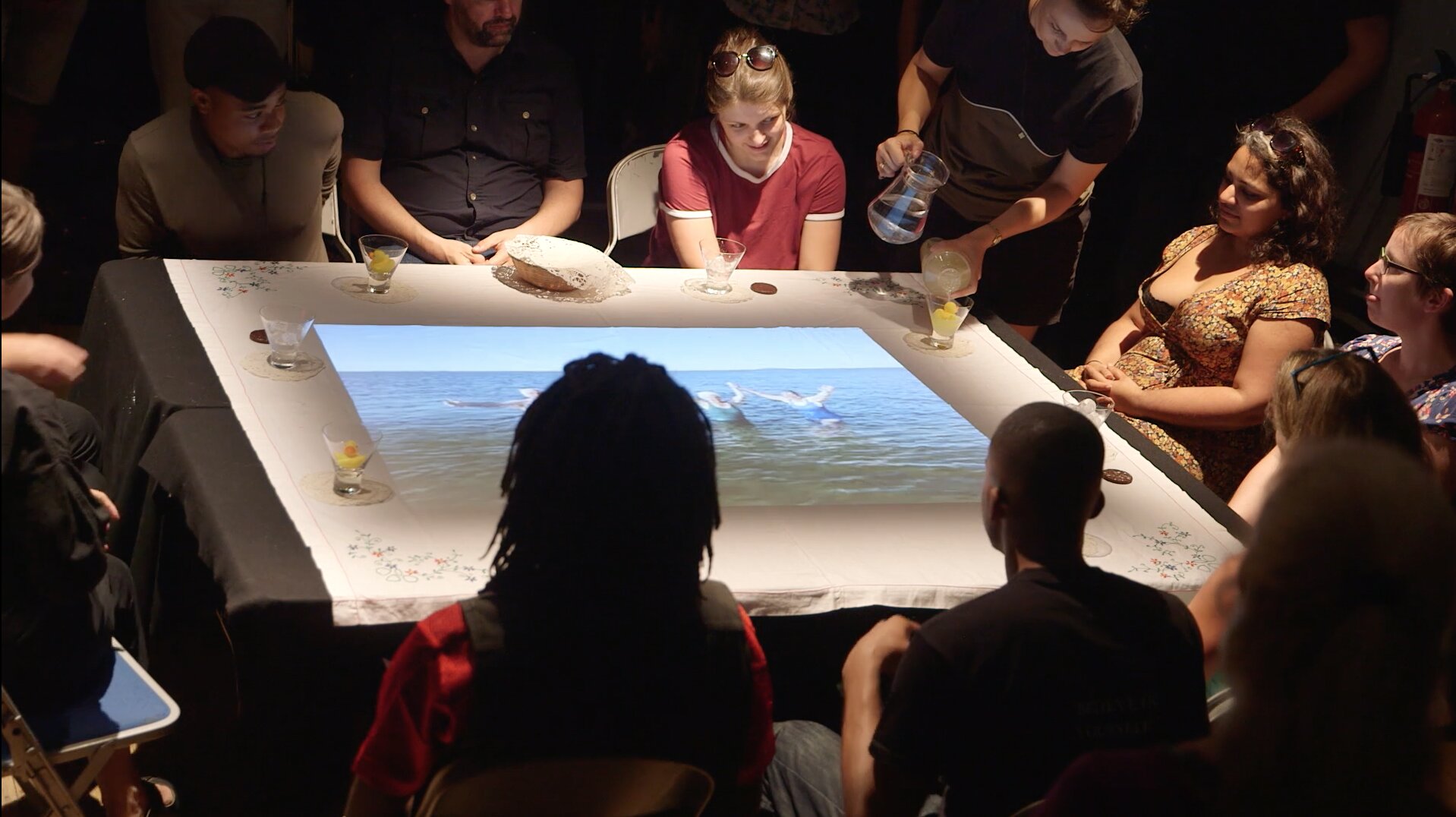 Participants gathered around a cloth-covered table with a projected seascape, pouring drinks during Rhiannon Armstrong's ritual performance