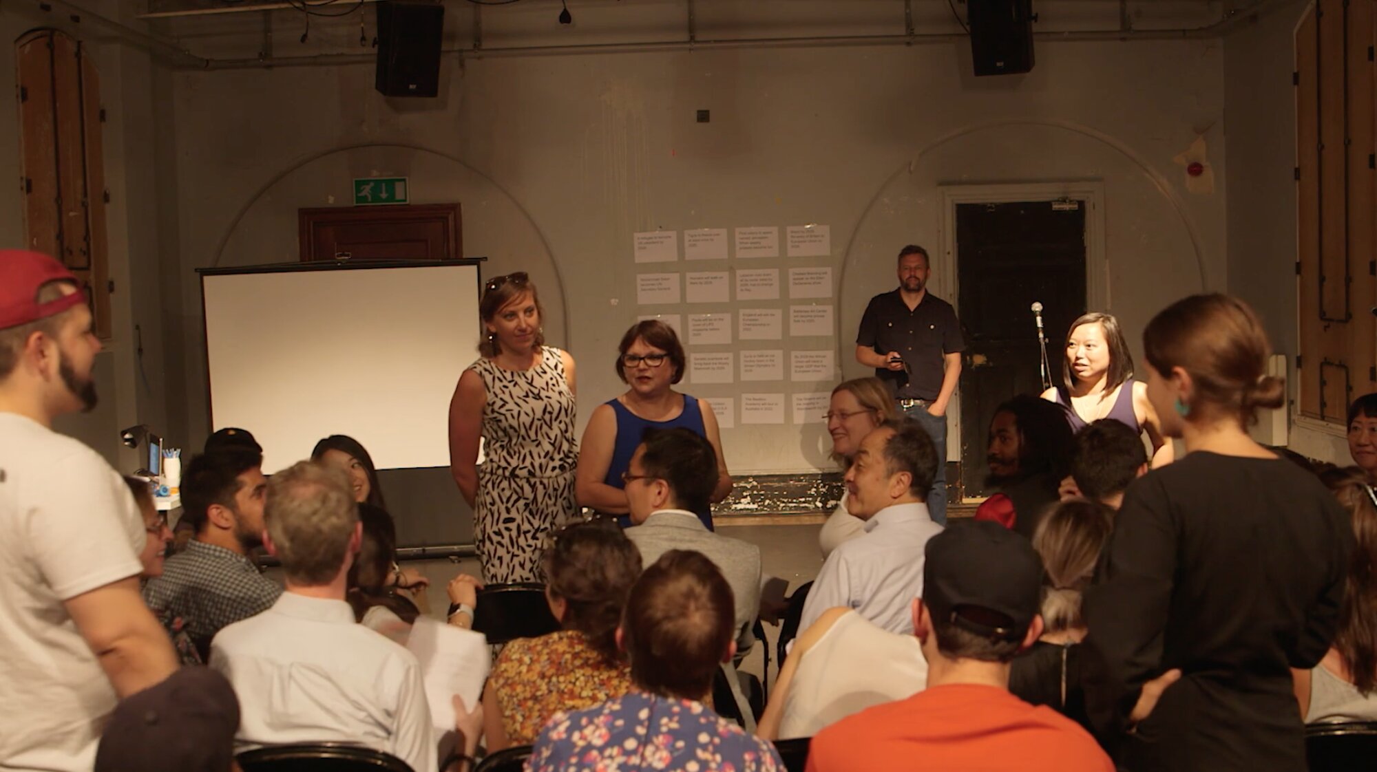 Audience members standing among seated participants as facilitators address the room during Tassos Stevens's game Ten
